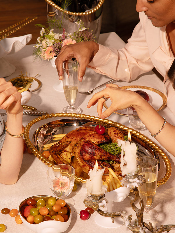 A close-up lifestyle shot of a dining table during a festive meal. A large gold-plated platter holds a roasted turkey with potatoes and green garnish, surrounded by lit white candles, glasses of champagne, and a bowl of colorful grapes.
