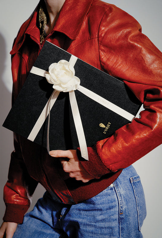 A person wearing a red leather jacket and blue jeans holds a black rectangular box with a large white fabric flower and white ribbon, against a plain light gray background.