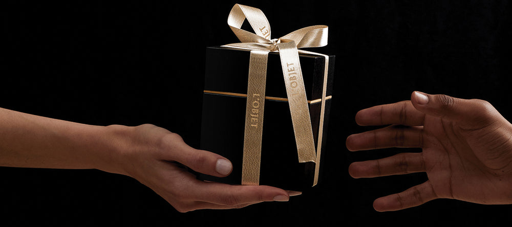 Two hands reaching towards a black gift box with a gold ribbon on a black background