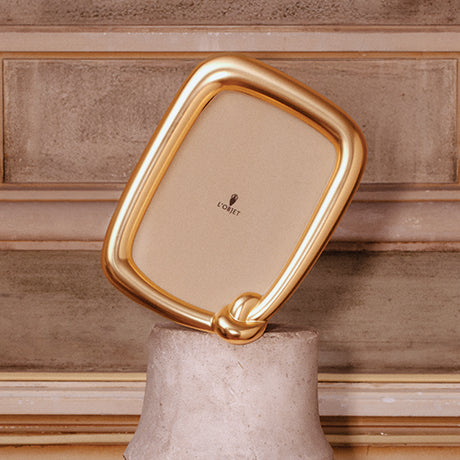 A gold-framed square mirror with a polished metallic finish and a decorative knot detail at the bottom, leaning against a textured beige stone wall on a stone pedestal. The brand name 'LOFFET' is printed in black on the mirror surface.