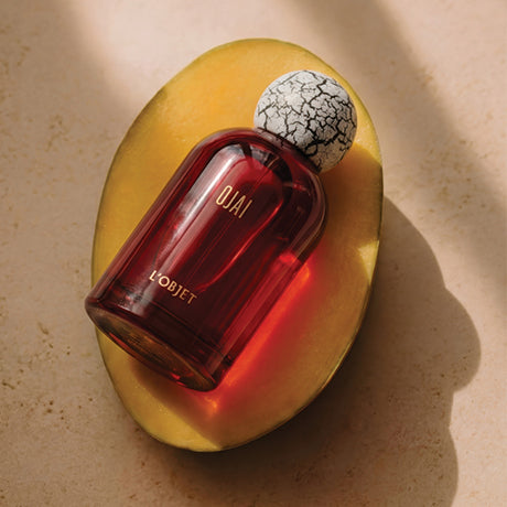 A bottle of OJAI L'OBJET perfume with a dark amber-red glass body and a white-and-black crackled stopper, resting on a sliced yellow mango against a textured beige background.