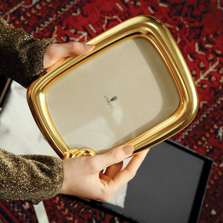 A person holds a square gold-framed mirror with a cream reflective surface, showing the 'LOVIT' brand logo. The background features a red patterned rug and a black gift box.