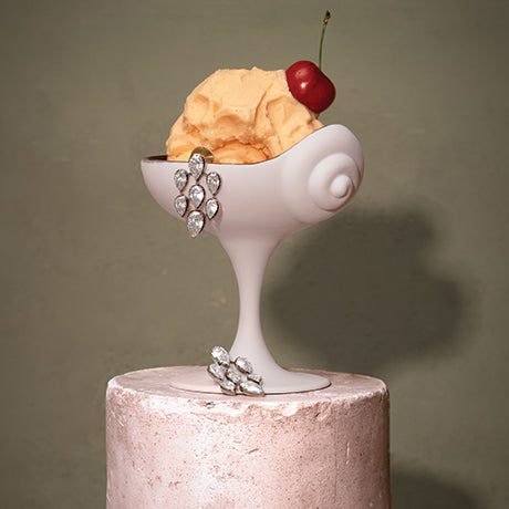 A sculptural white ceramic dessert bowl or pedestal dish holding a scoop of orange ice cream topped with a red cherry, displayed on a pale pink cylindrical pedestal against a muted olive-gray background.