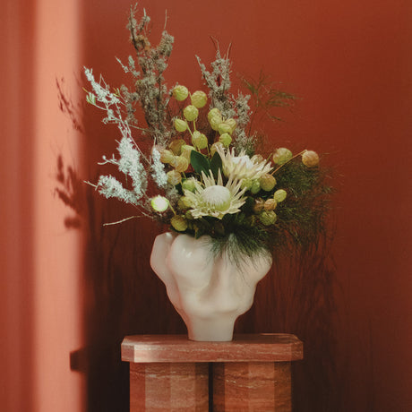 Floral arrangement in a white ceramic head vase on a wooden pedestal against a red wall.
