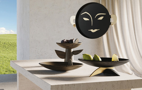 Surreal tabletop in a minimalist space set with L'Objet Kelly Behun scultural wooden serving bowls. A black resin and brass serving tray with illustrated face and pirced brass ear handles suspends above. 