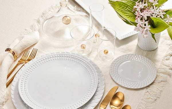 Table setting with Aegean white charger plate with wave motif border, Perlee white porcelain dinner plate, Perlee white vase and Oro pitcher and glasses.