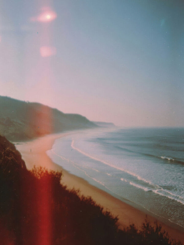 A scenic coastal landscape photograph showing a sandy beach curving along the ocean, with dark cliffs in the foreground and a hazy blue sky above. The image has a vintage, filtered look with soft light and lens flare.