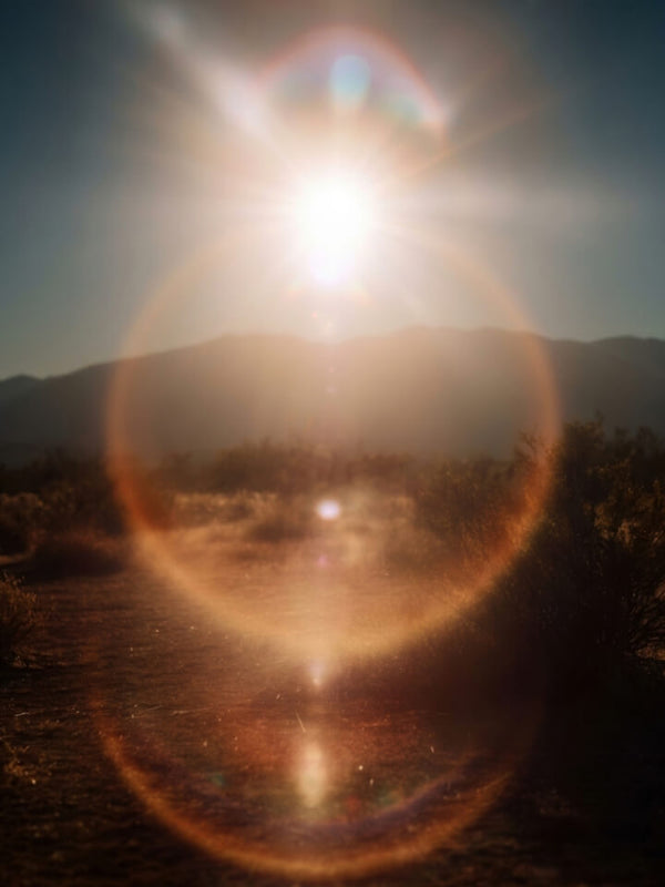 A scenic landscape photograph of a sunlit desert path with mountains in the background, heavily overlaid with a large circular lens flare and bright sun glare.