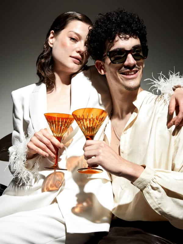 A man and a woman pose closely together while holding orange faceted cocktail glasses. The woman wears a white blazer with white feather trim, and the man wears a cream shirt and dark sunglasses.