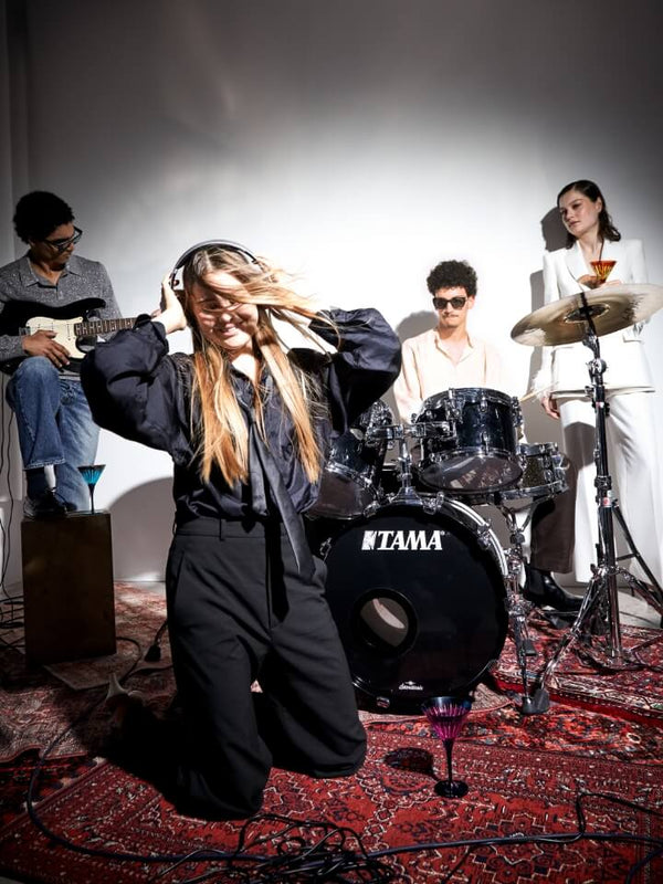 A band performance scene with a woman in the foreground kneeling and holding a microphone, surrounded by guitar, drums, and cymbal players. Visible product branding includes TITAM on the drum kit.