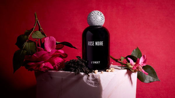 Shop Fragrance Collection. Discover Rose Noire Fragrance. Black Porcelain Bottle with cracked wood top. On a chipped stone pedestal with roses and other aromatics around it. 