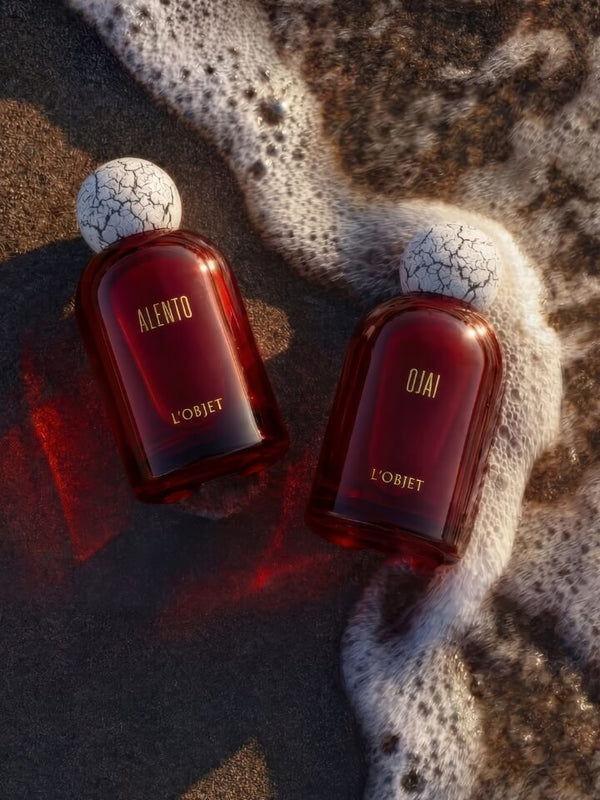 Two L'Objet Alento and Ojai perfume bottles with dark red glass and white textured caps, placed on wet sand with foamy ocean water around them.
