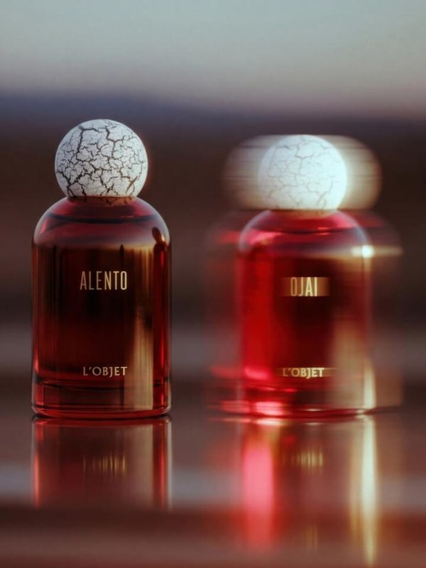 Two L'Objet Alento perfume bottles with deep red glass and white cracked-embroidered spherical caps, displayed on a reflective surface with a soft blurred background.
