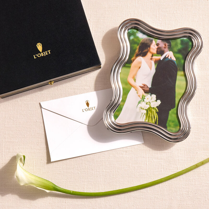 Decorative photo frame with a couple and flowers, next to L'OBJET branded items on a beige surface.