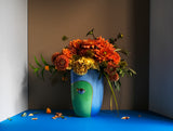 L'OBJET x LITO Eye Motif Vase in Green and Blue with orange and yellow flowers arranged on a sky blue tabletop and brown wall.