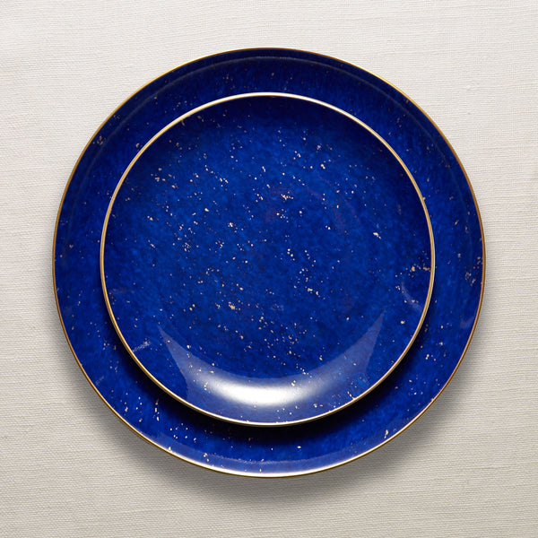 Lapis blue porcelain dinnerware stack with 24K gold rim and speckled accents. On top of a beige woven tablecloth.